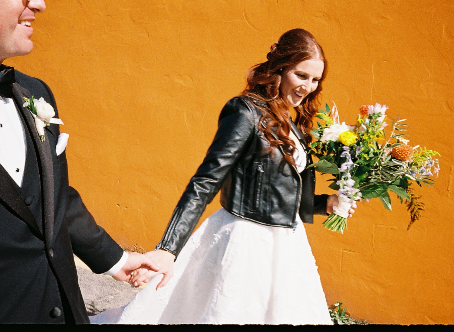 Lauren and Adam walking hand in hand with a bouquet against an orange wall